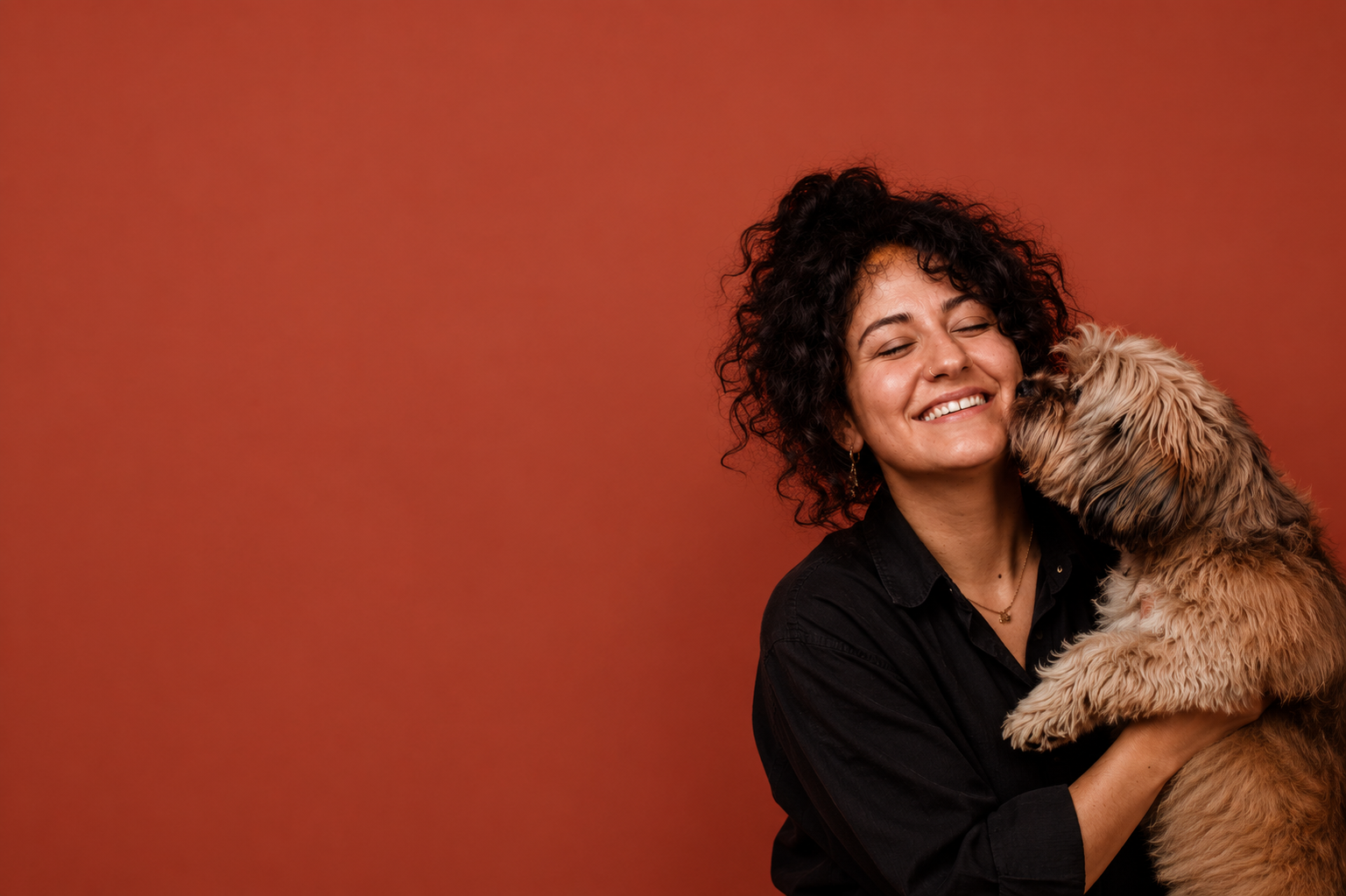 Smiling woman holding a small fluffy dog as it nuzzles her cheek against a warm burnt orange studio background.